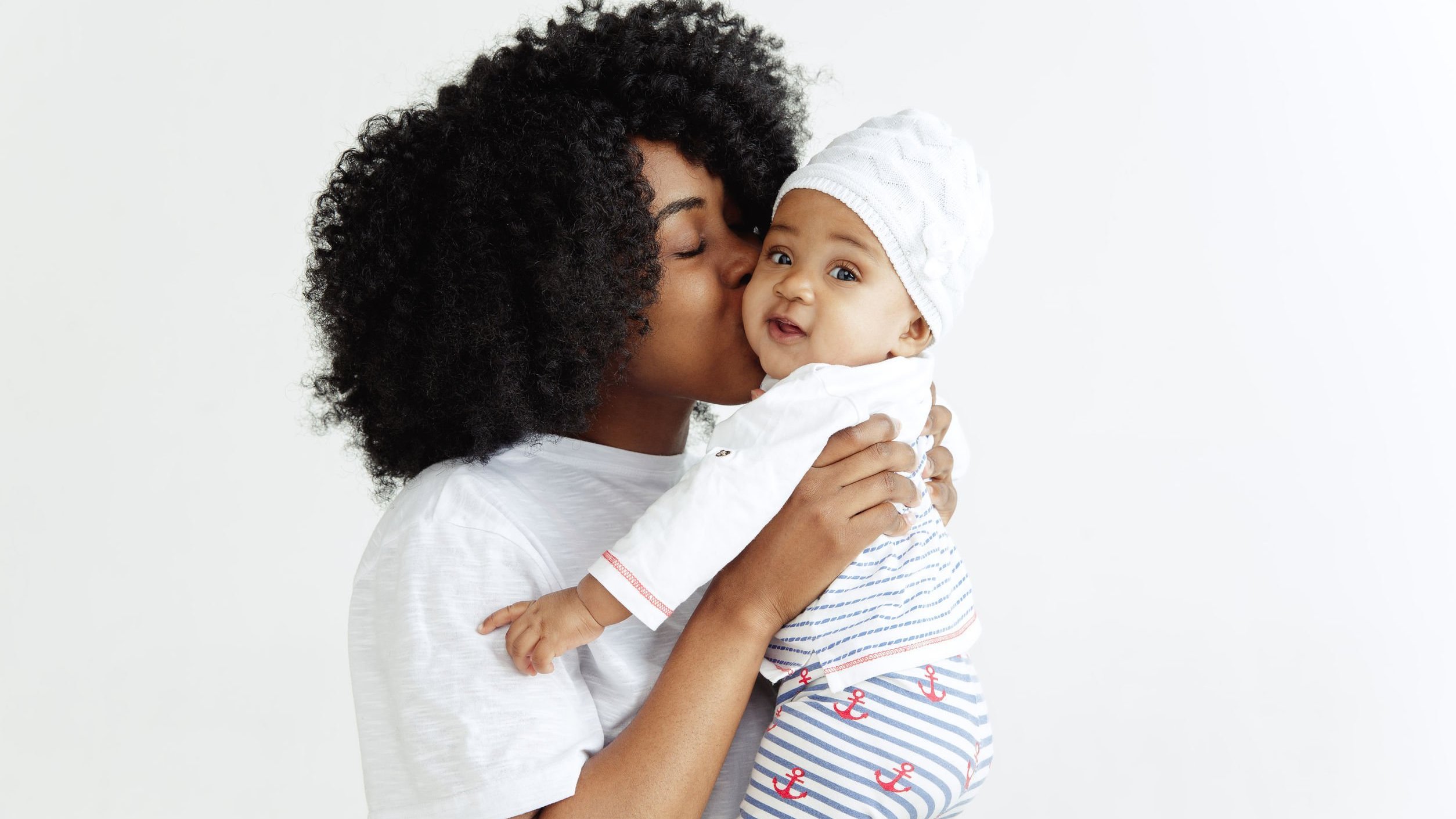 Mulher negra de cabelos curtos e crespos, segurando bebê negro e beijando-o na bochecha.