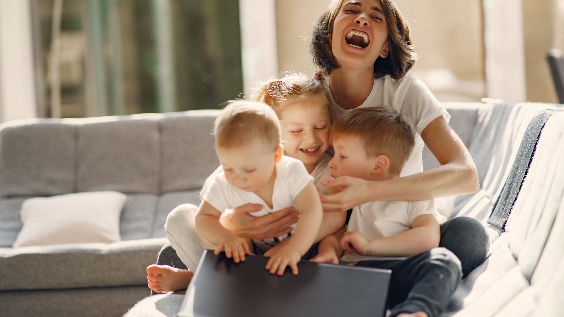 Imagem de uma mãe feliz, sentada no sofá. No seu colo, estão sentados os seus três filhos.