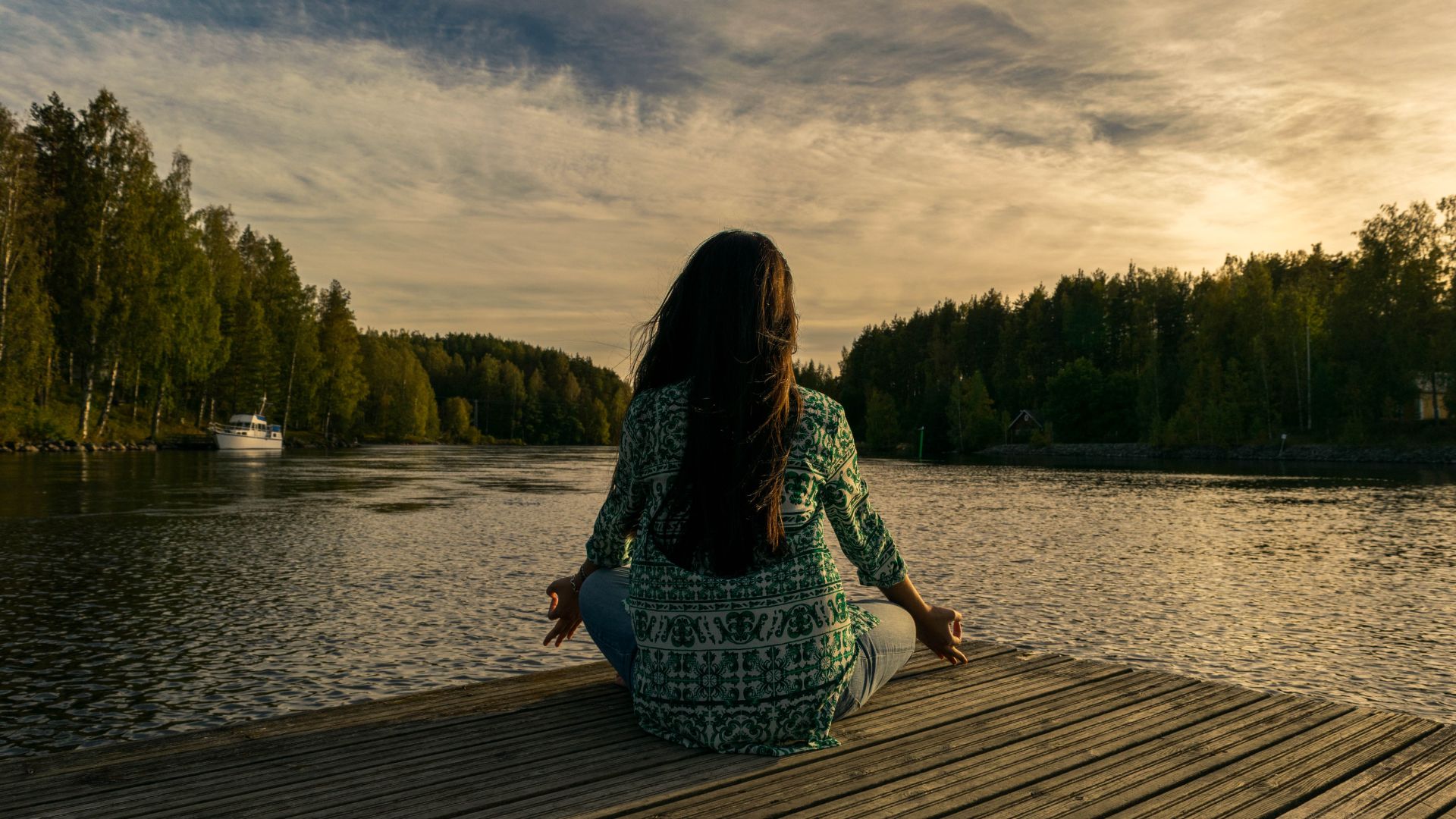 Imagem de uma mulher de costas, sentada em posição de meditação e uma paisagem de lago e natureza ao seu redor.
