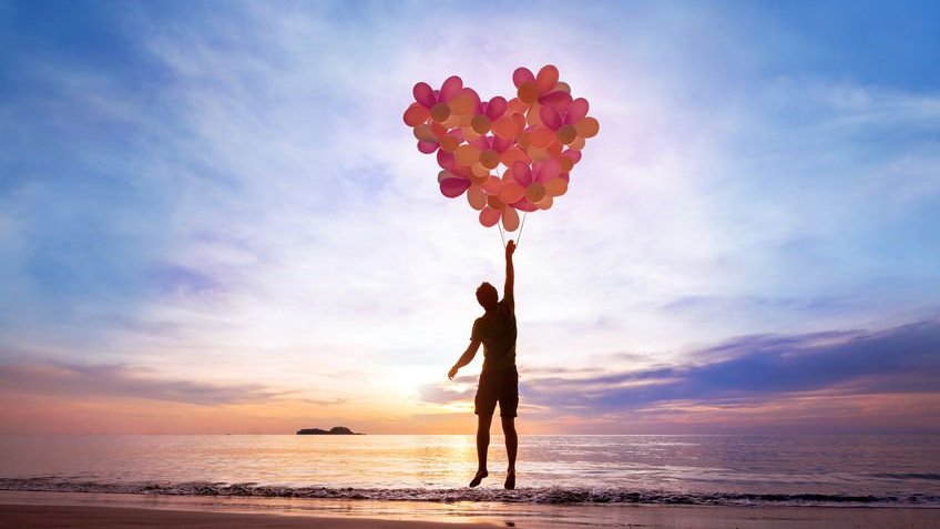 Silhueta de homem segurando balões formando coração com fundo de praia e sol se pondo