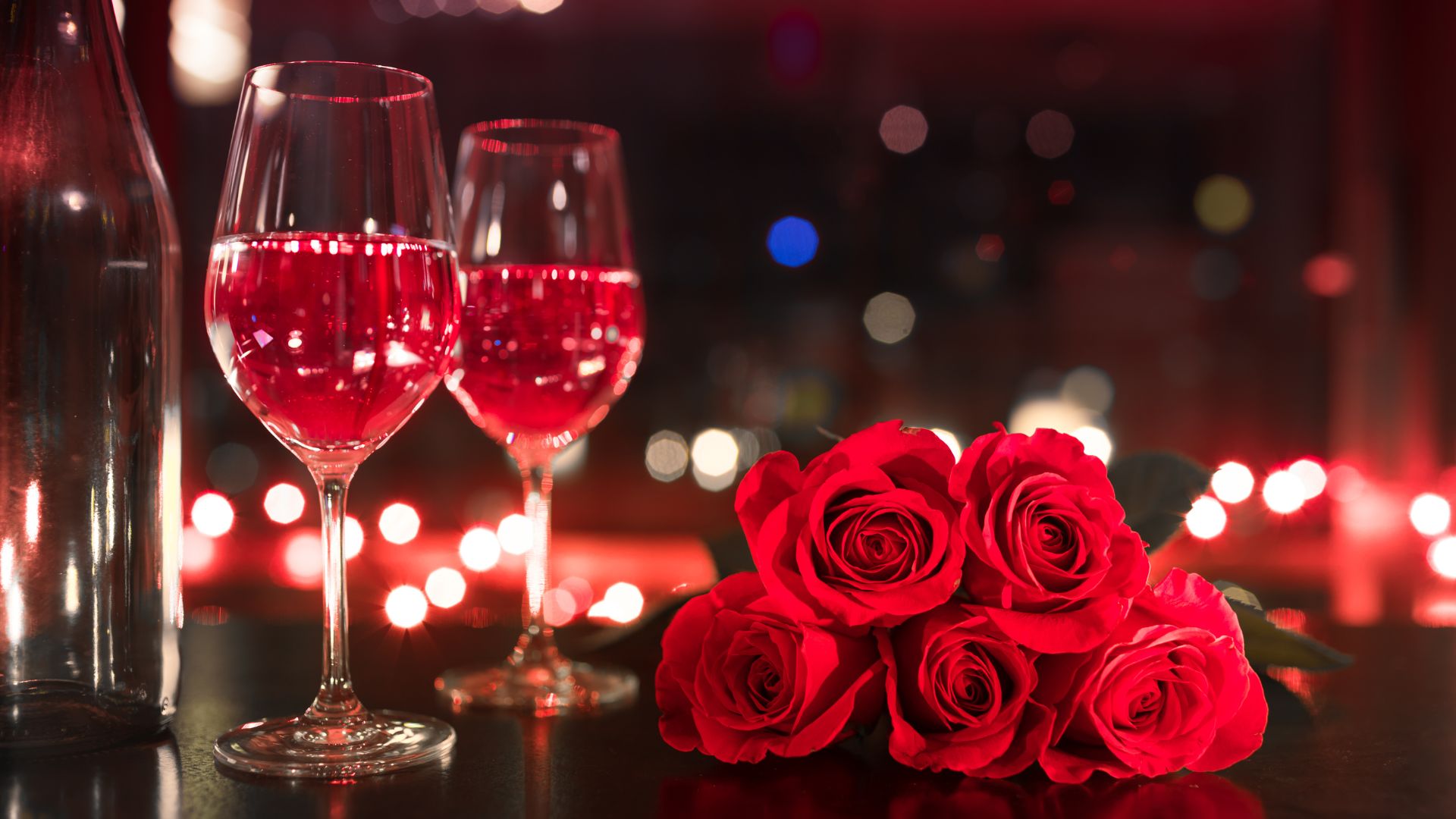 Imagem de duas taças de vinho, uma garrafa ao lado e rosas vermelhas na mesa.