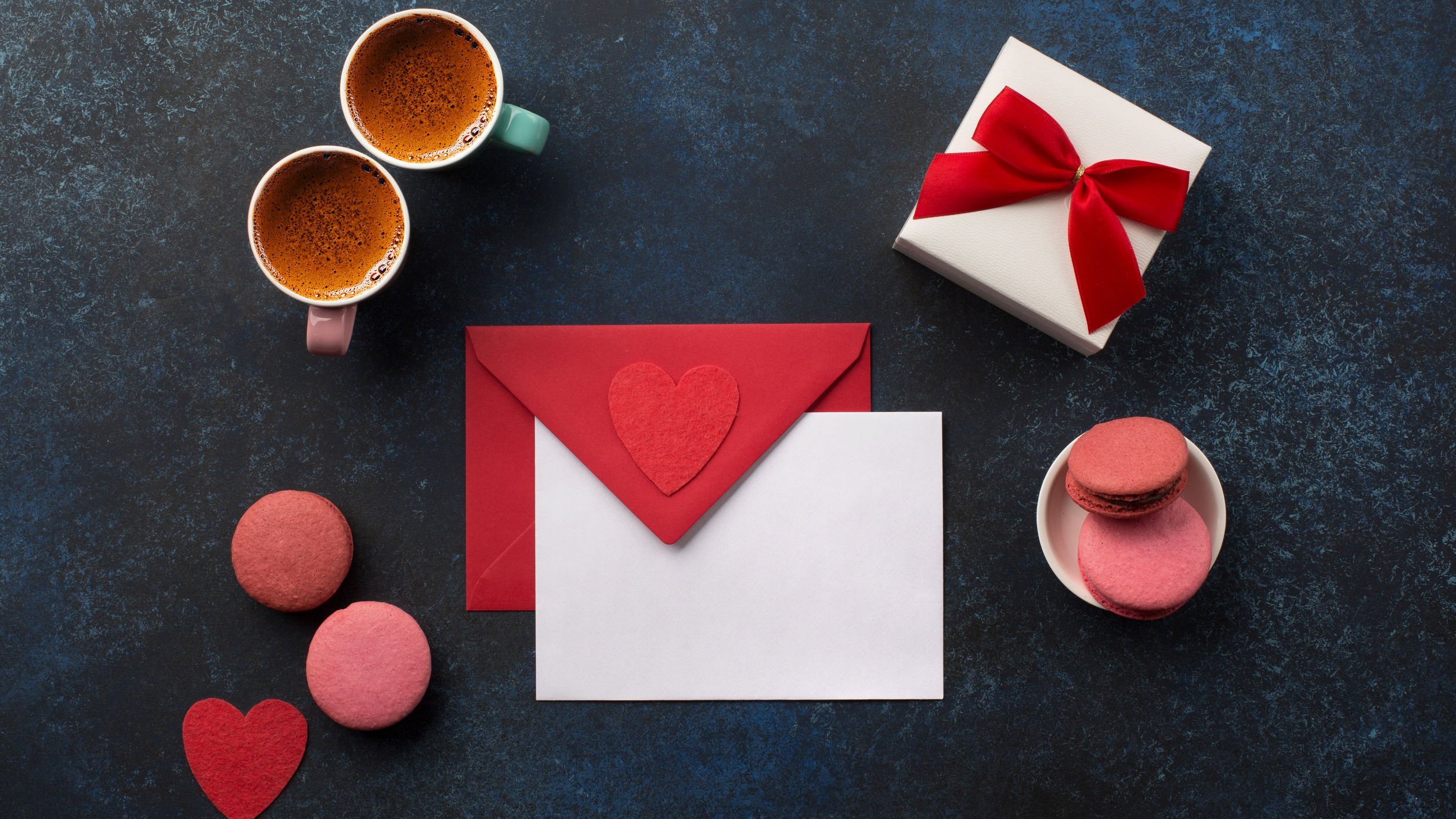 Duas canecas com café, uma caixa de presente com laço e um envelope vermelho com um coração e um papel branco na frente.