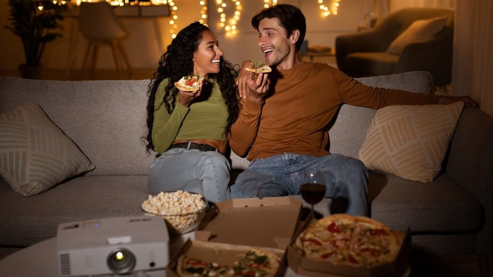 Casal sentado em um sofá comendo pizzas enquanto projetam um filme para ver