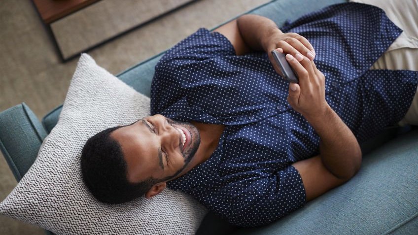 Homem deitado no sofá mexendo no celular sorrindo visto de cima