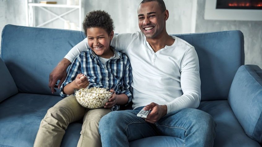 Pai e filho comendo pipoca enquanto assistem a um filme, no sofá.