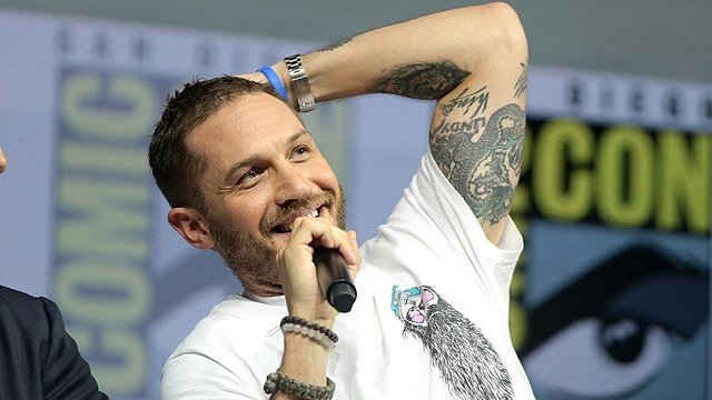 Tom Hardy sorrindo em evento, segurando um microfone com a mão direita enquanto tem sua mão esquerda em sua cabeça.