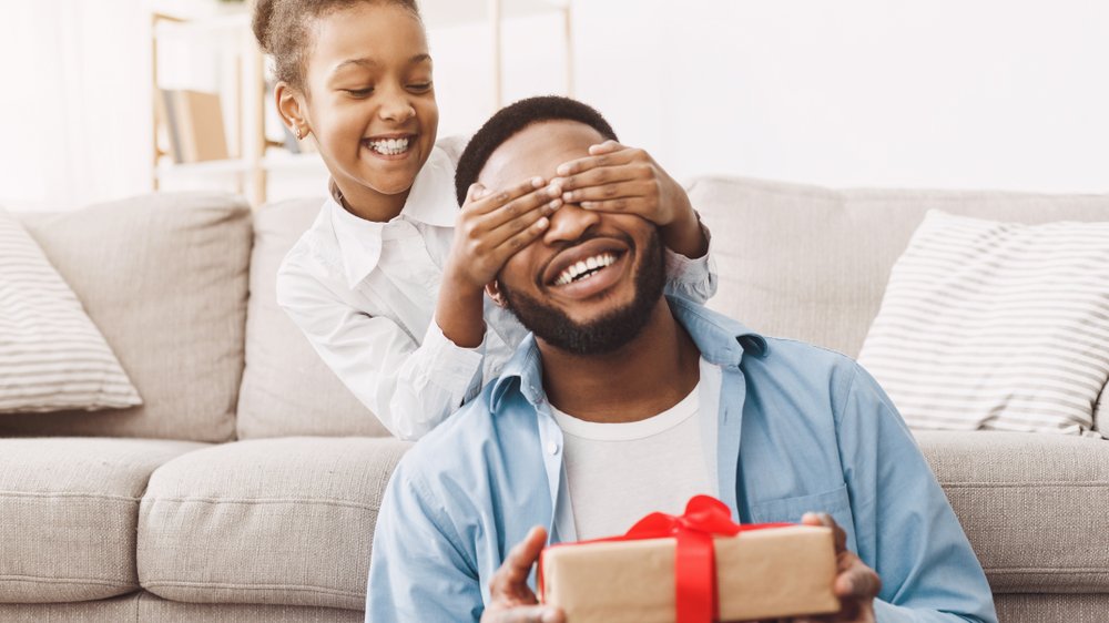 Filha tampando o olho do pai que segura um pacote de presente.