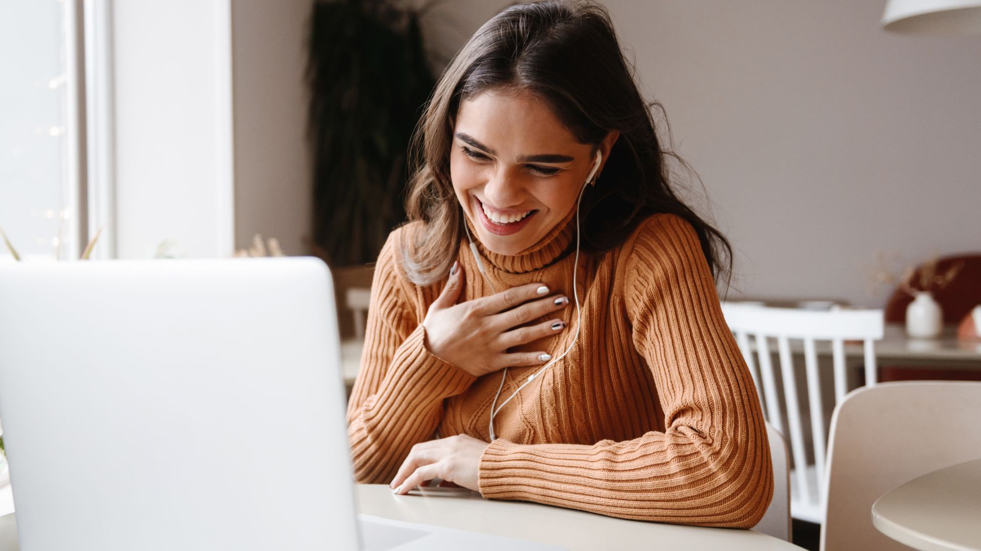 Imagem de uma mulher na frente do computador sorrindo com a mão no coração, como se estivesse em uma chamada de vídeo.