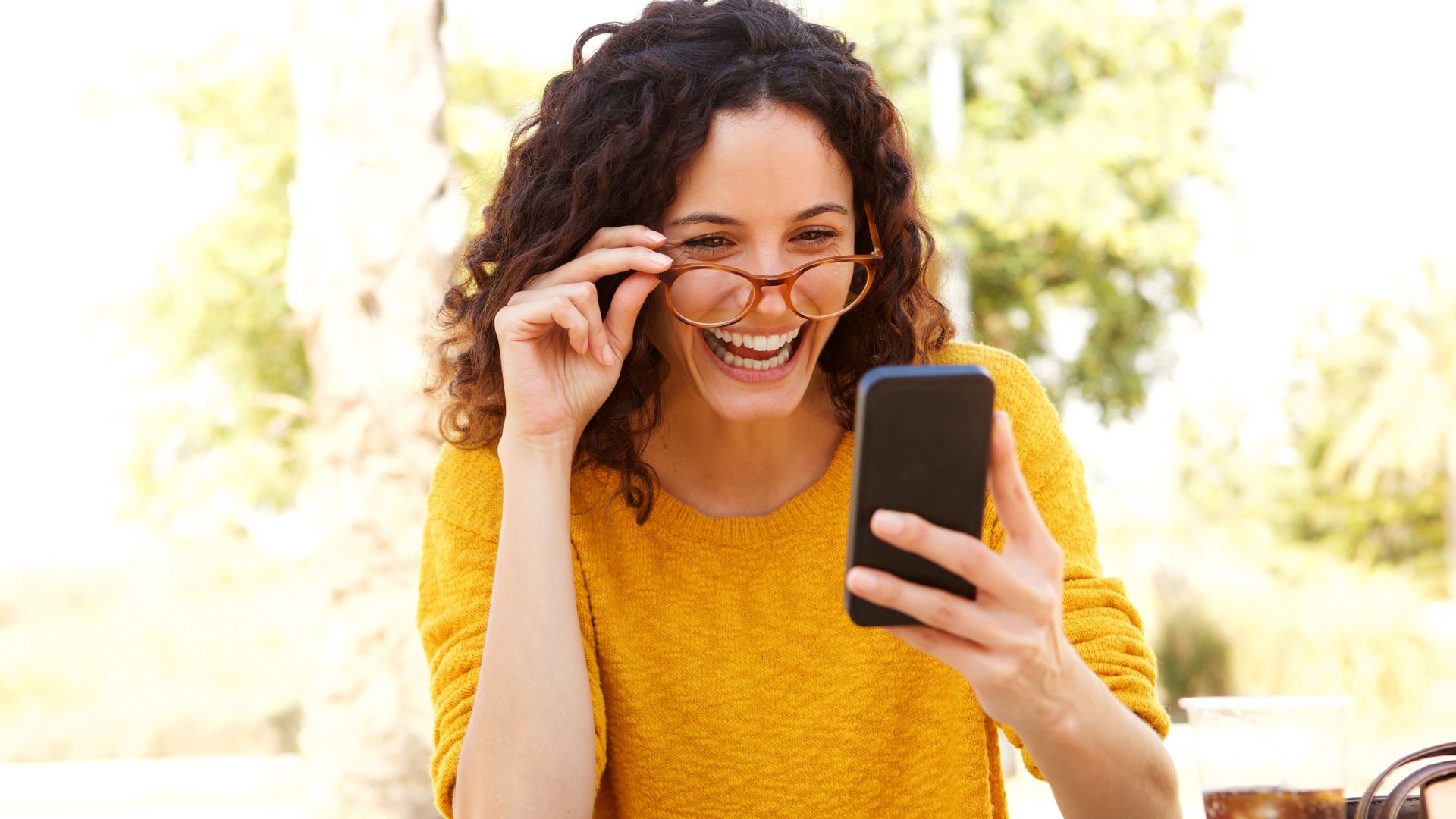Mulher de óculos com roupa amarela, olhando o celular e dando risada