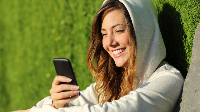 Menina adolescente usando um celular em um parque com fundo verde.