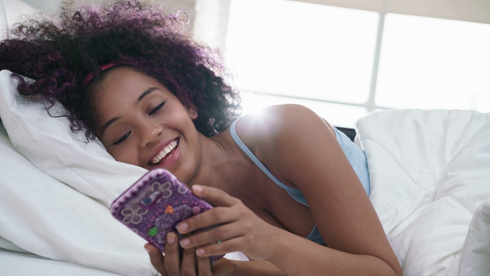 Mulher negra deitada numa cama, segurando um celular e com expressão risonha.