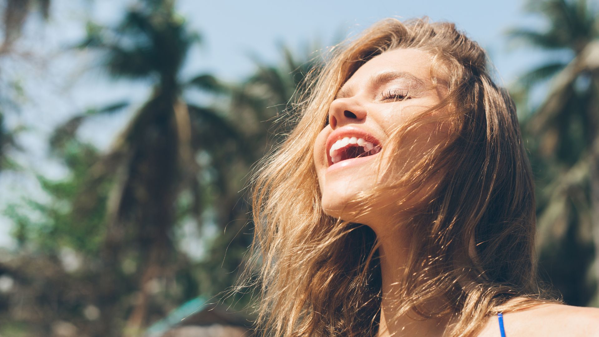 Imagem de uma mulher de cabelos longos, sorridente e feliz. Ao fundo da foto, alguns coqueiros.