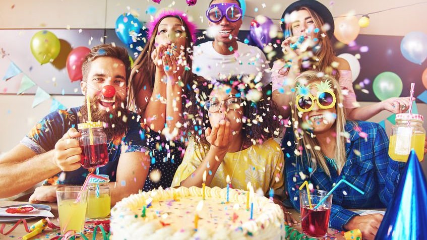 Grupo de seis pessoas em frente a um grande bolo coberto de glacê branco cercado por confetes na sala comemorando aniversário.