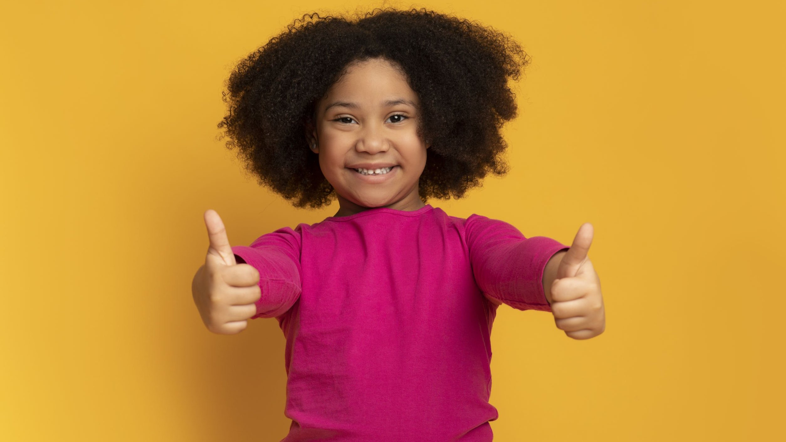 Menina com os braços esticados para a frente, e os dois polegares erguidos para cima, com os dedos fechados.