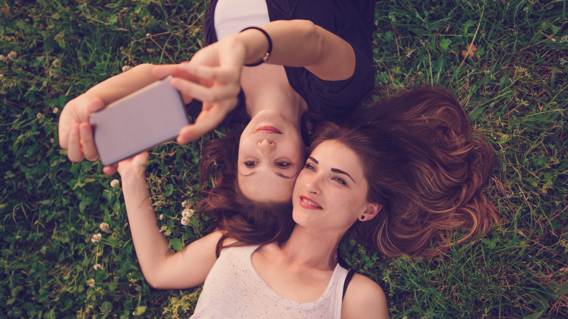Duas amigas deitadas na grama tirando uma foto pelo celular