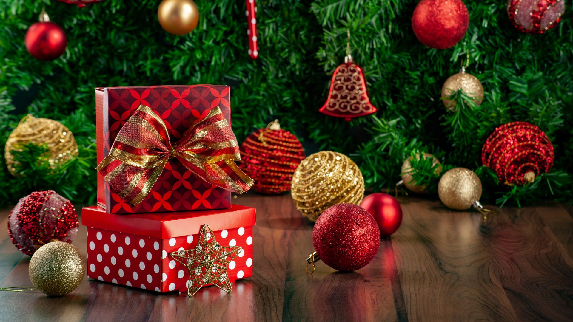 Imagem contendo ao fundo um grande pinheiro de Natal decorado com bolas e outros enfeites nas cores vermelho e dourado. Em destaque duas caixas de presentes decoaradas com laço.