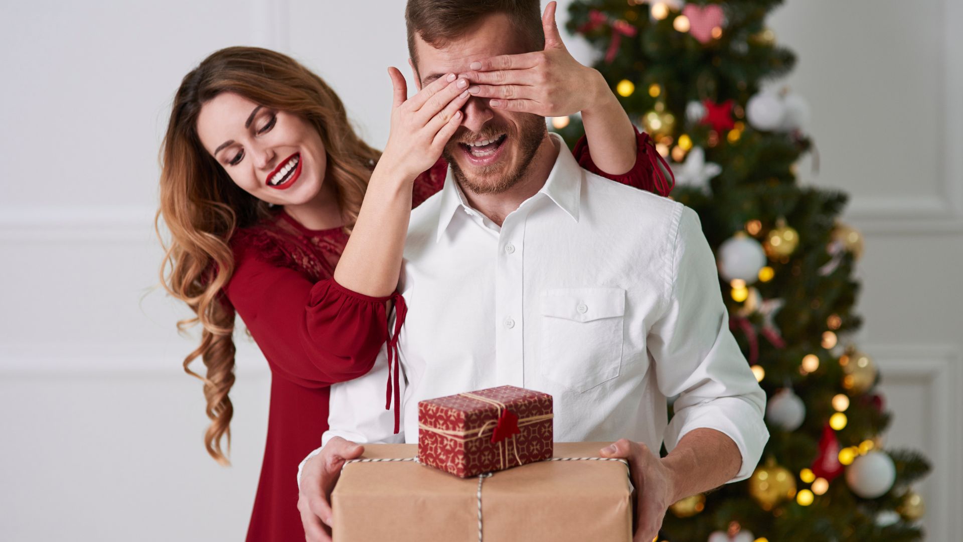 Imagem de um jovem casal. Ela usa um vestido vermelho. Ele usa uma camisa branca. Ela está tampando os olhos dele e entregando duas caixas de presente. Ao fundo uma linda árvore de Natal.
