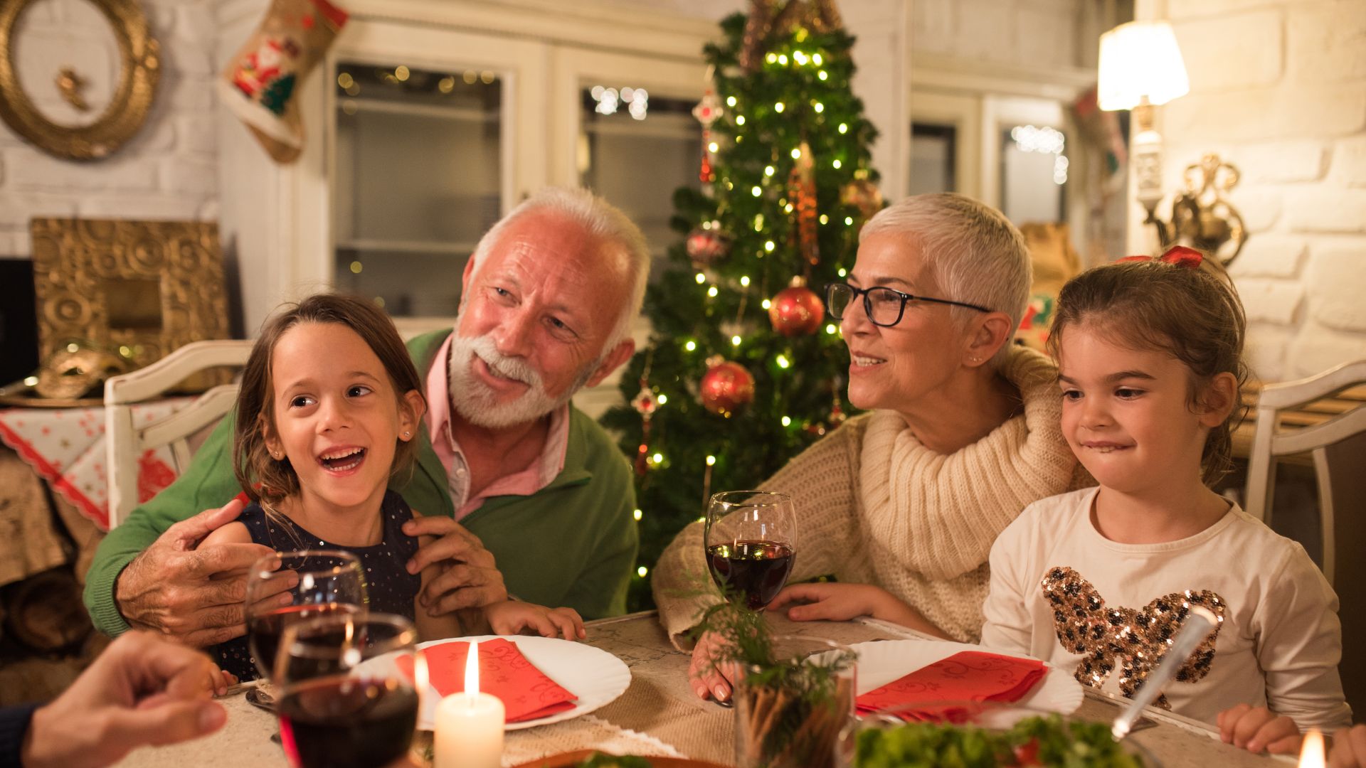 Imagem de um casal de idosos, acompanhados de suas netinhas. Todos estão sentados à uma mesa decorada com enfeites natalinos. Ao fundo, uma linda árvore de Natal.