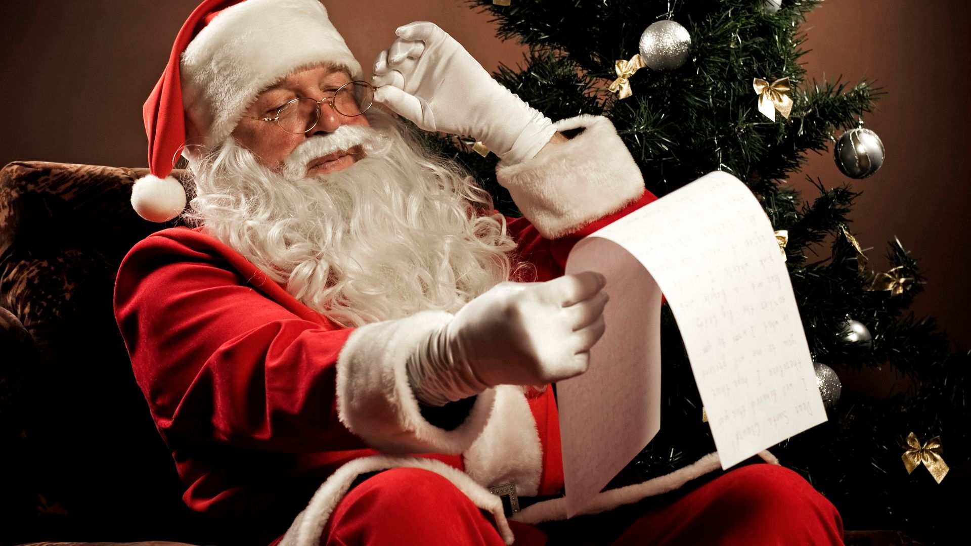 D-Keine de Getty Images Signature para o Canva Imagem de um papai noel sentando em uma poltrona aveludada na cor marrom. Ele lê uma carta e ao lado dele temos uma árvore decorada.