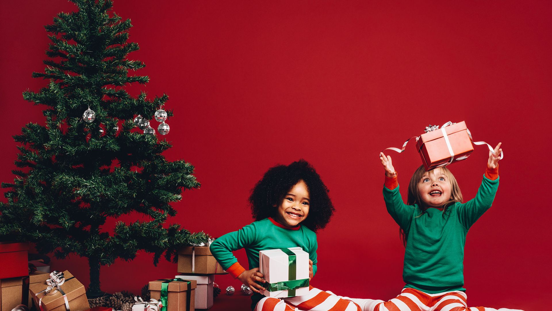 Imagem de fundo vermelho. Do lado esquerdo da tela um pinheiro de Natal e sob ele, caixas de presentes no chão. Em destaque, duas crianças vestidas com calças listradas vermelho e branco e blusa de manga longa na cor verde. Ambas estão felizes e seguram seus presentes.