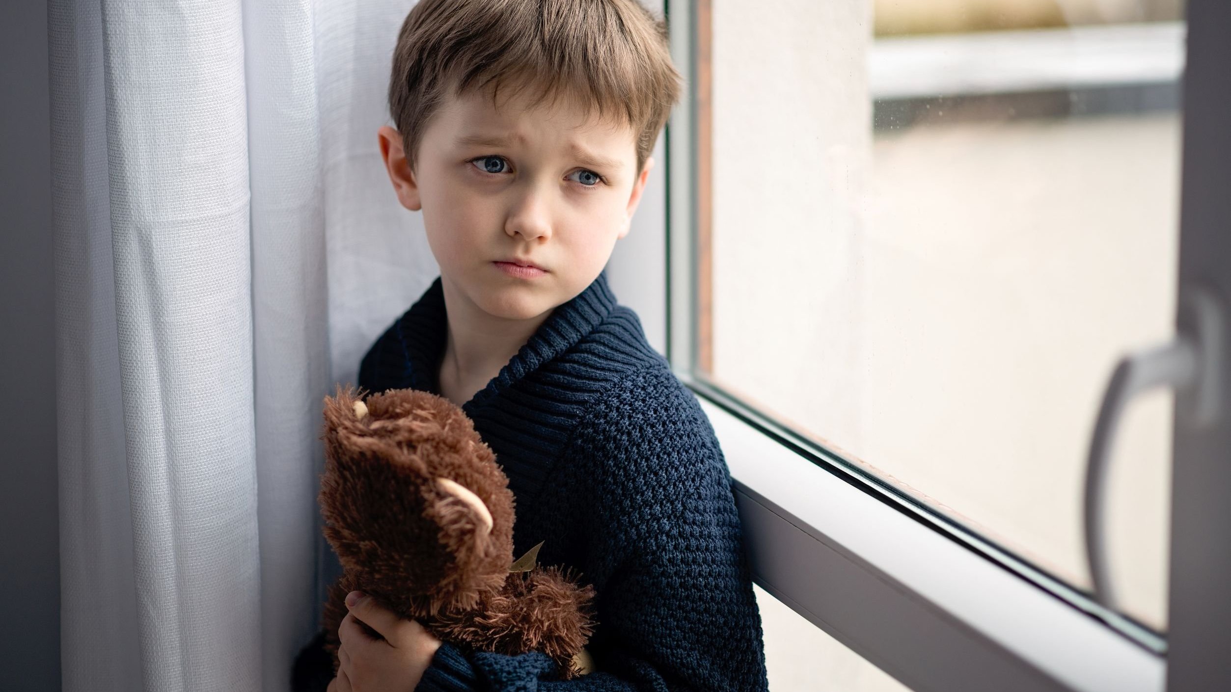 Criança triste olhando para o lado da janela, segurando ursinho de pelúcia