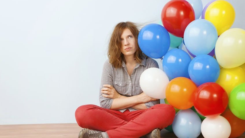 Mulher sentada no chão com expressão de tédio ao lado de balões coloridos.