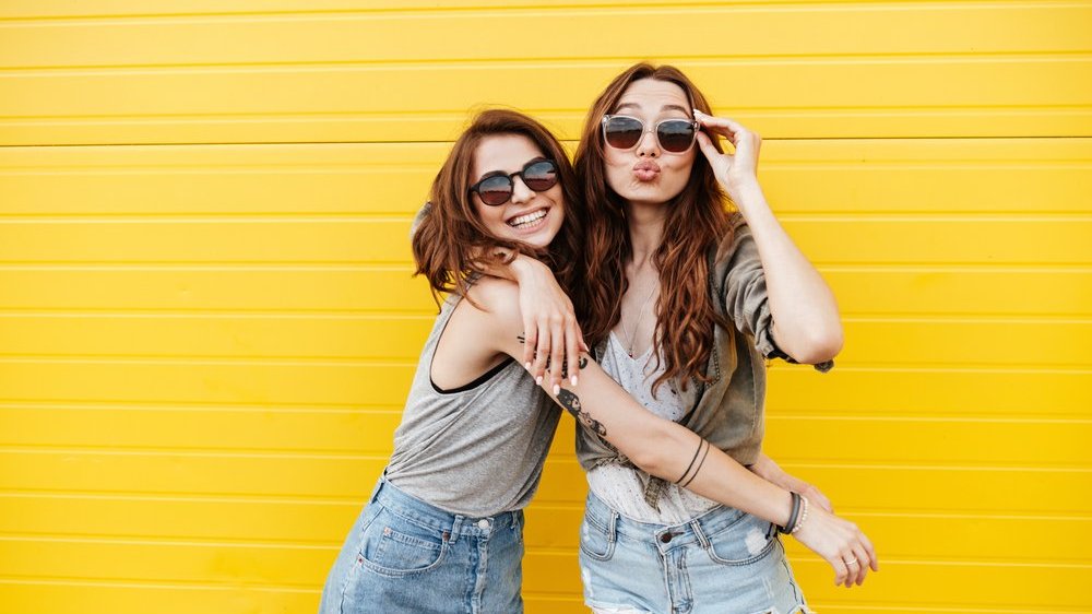 Duas amigas abraçadas usando óculos de sol, uma está sorrindo e a outra mandando beijo