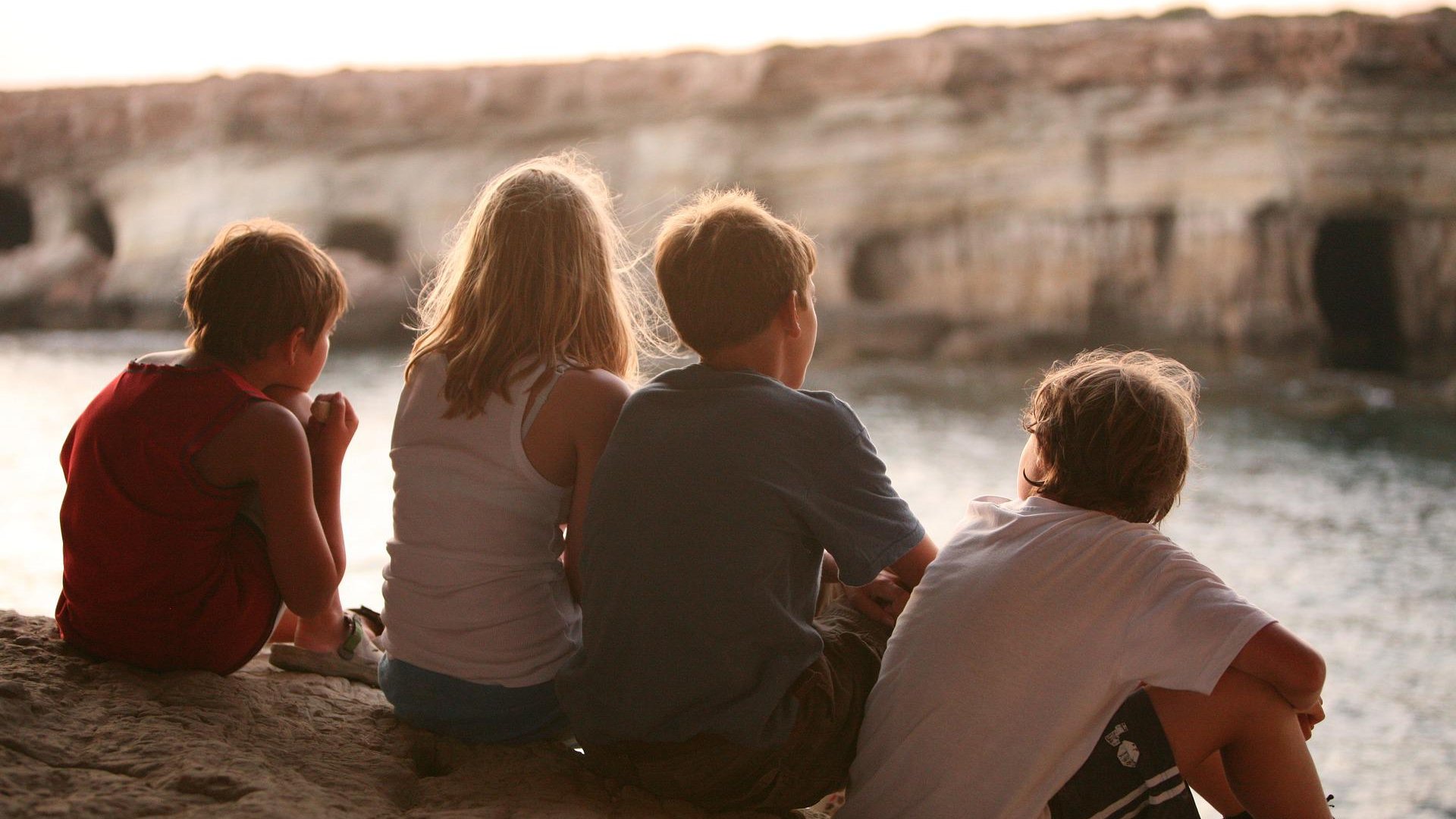 Quatro crianças sentadas de costas em pedreira observando lago.