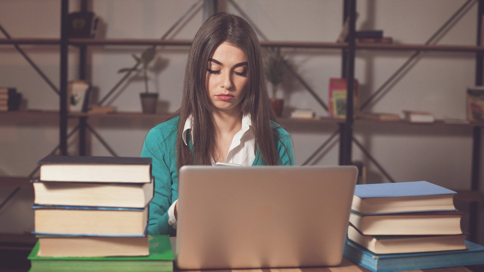 Mulher branca em frente à computador e rodeada de livros.