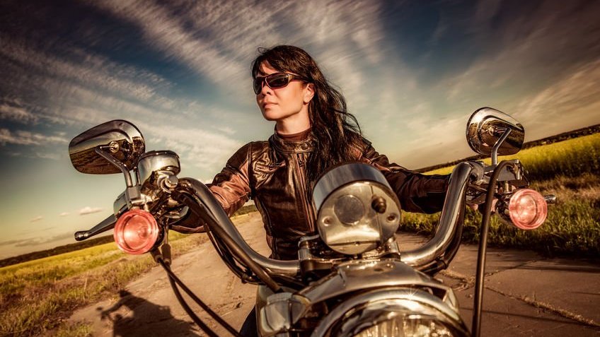 Mulher usando jaqueta de couro e óculos escuro pilota uma moto em um deserto.