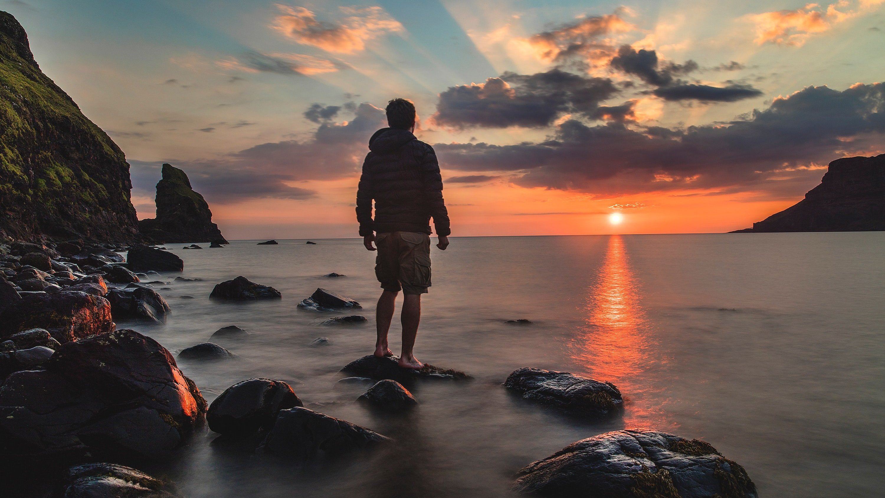 Homem em pé sobre uma rocha na praia, olhando para o mar, sob o pôr do sol.