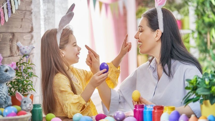 Mulher e menina, ambas usando tiaras de orelhas de coelho, pintando ovos, e as duas ameaçando a pintar o nariz da outra.