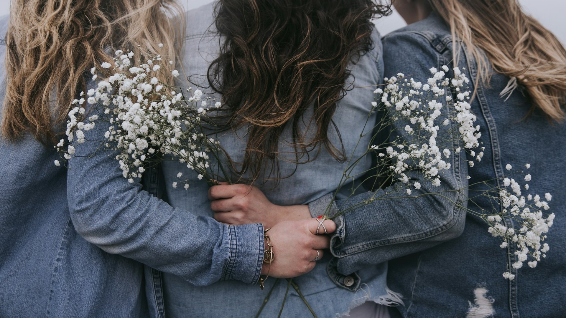 Três mulheres de costas, vestindo jaquetas jeans. Elas estão se abraçando e segurando pequenos ramos de flores.