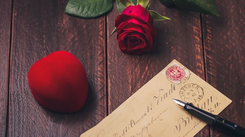 Um estojo de anel em veludo vermelho e formato de coração, uma carta e uma caneta sobre uma mesa de madeira.