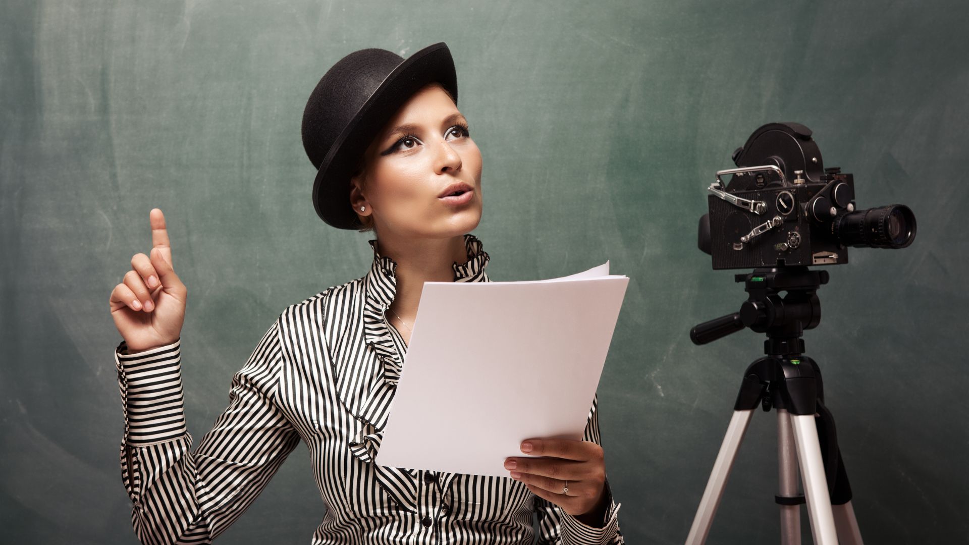 Imagem de uma atriz usando uma camisa listrada nas cores branco e preto e um chapéu preto, ensaiando o seu roteiro. Ao lado, uma câmera de TV.
