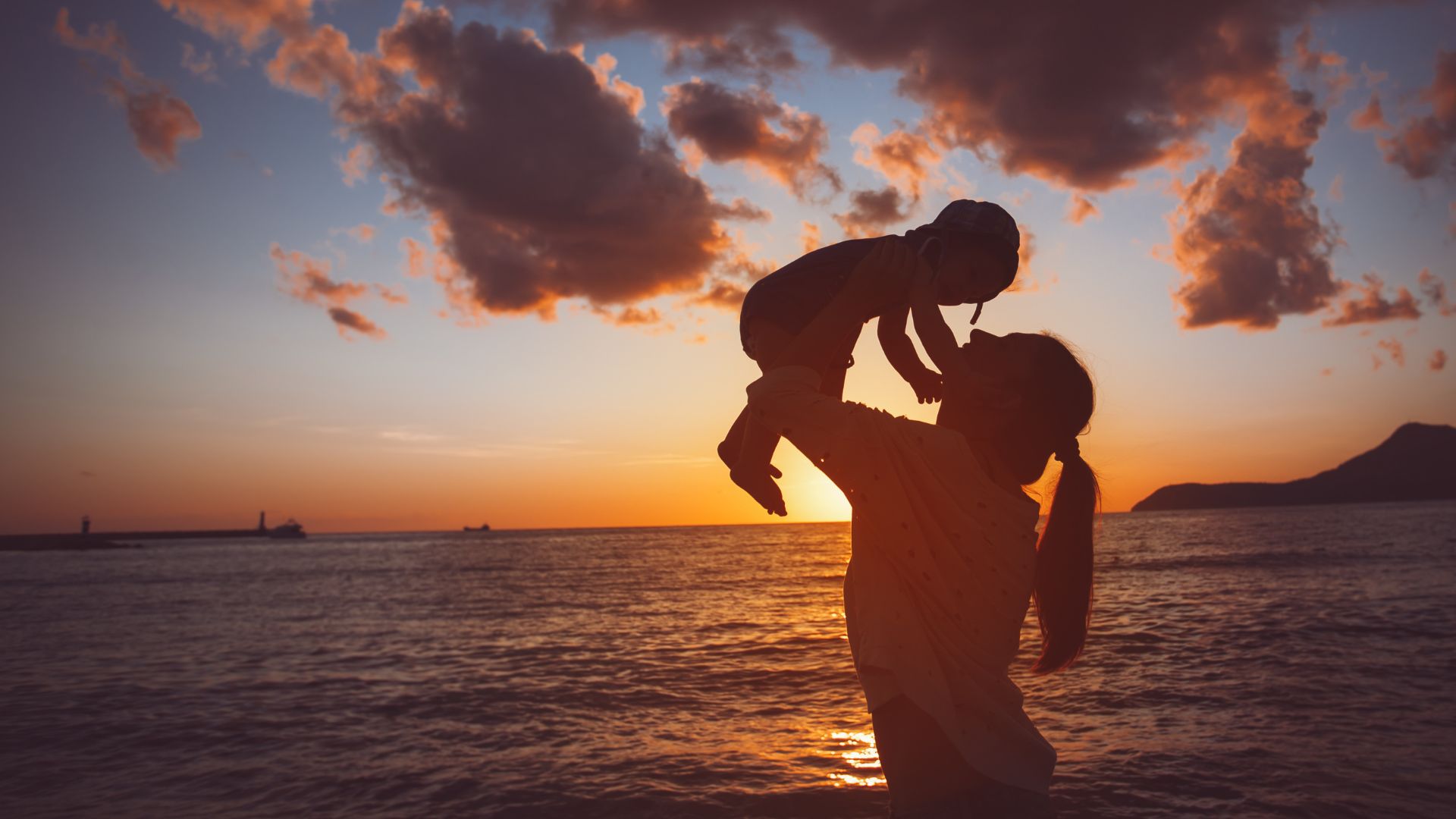 Imagem do mar e ao fundo o pôr do Sol. Em destaque, a mãe segurando no colo o seu filho.