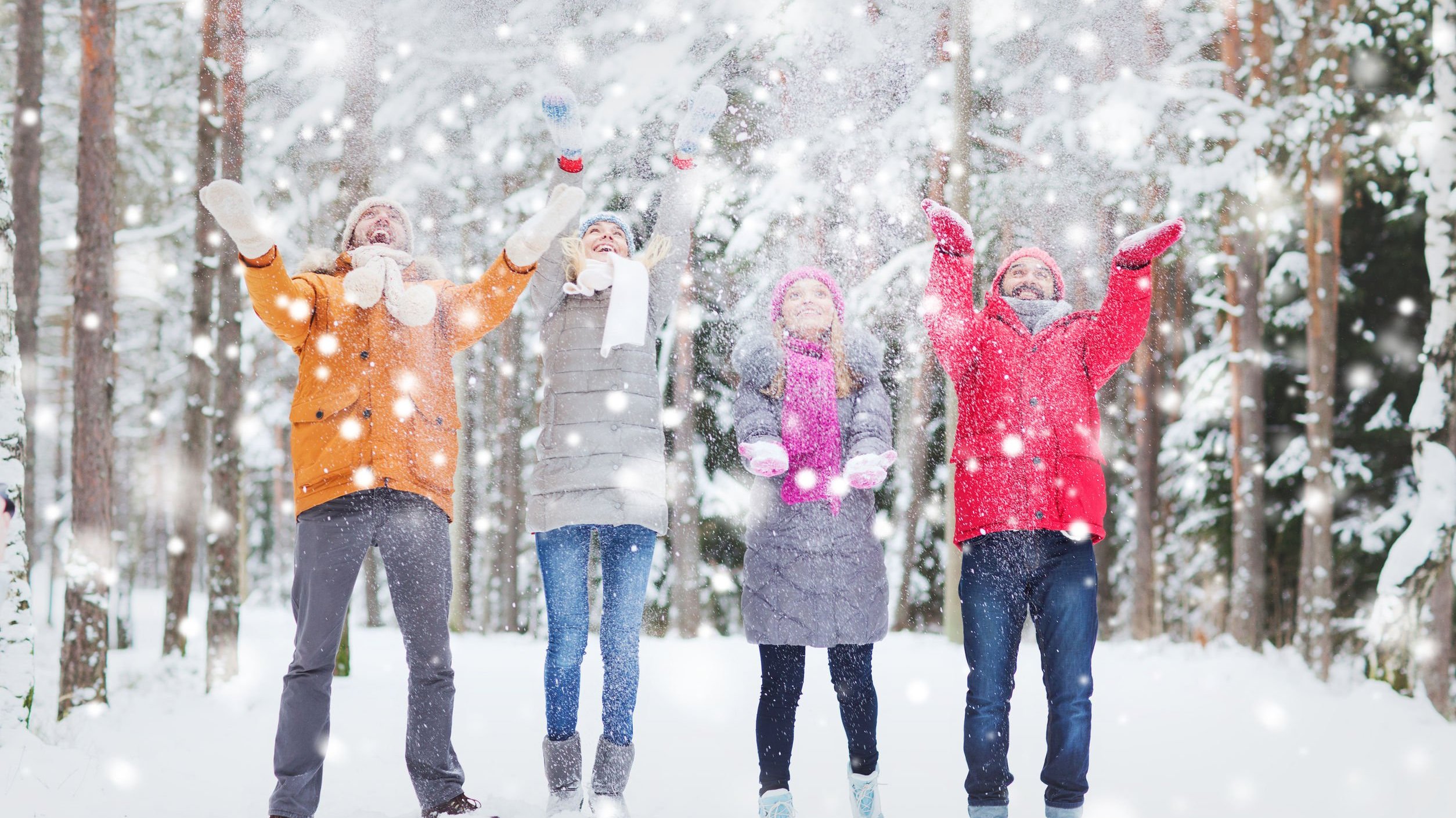 Quatro pessoas agasalhadas brincam com flocos de neve em ambiente exterior. Elas estão com os braços erguidos e sorriem.