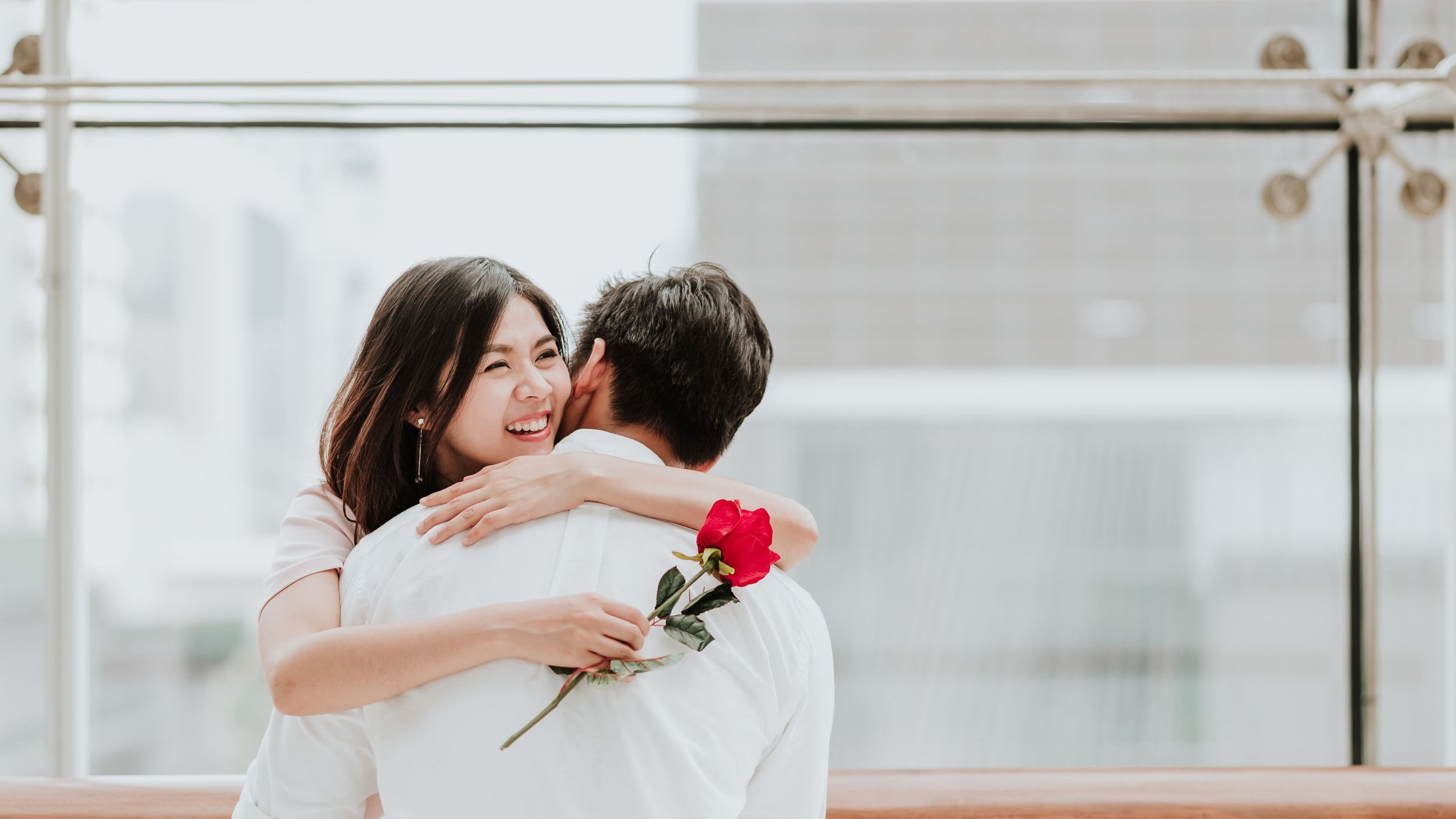 Mulher asiática e homem brancos se abraçando, a mulher segurando uma rosa vermelha e com expressão sorridente