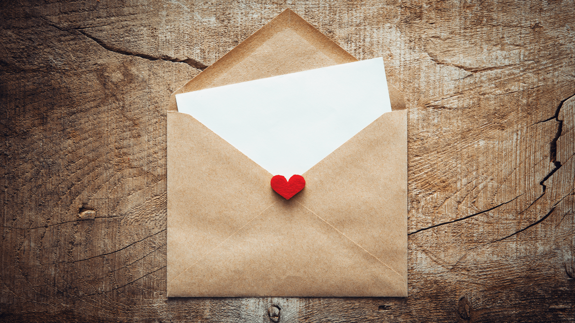 Imagem de um envelope de papel cartão com uma carta saindo dele e o selo de coração, em uma mesa de madeira.