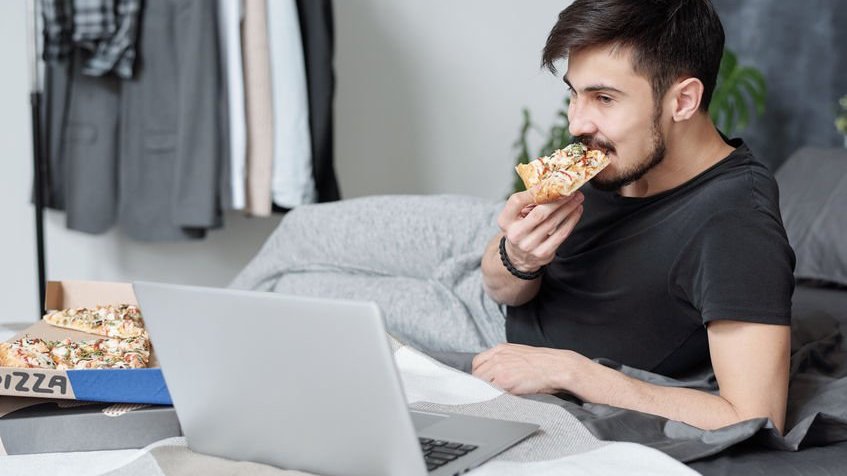 Homem deitado em cama, comendo pizza, enquanto assiste algo em notebook.