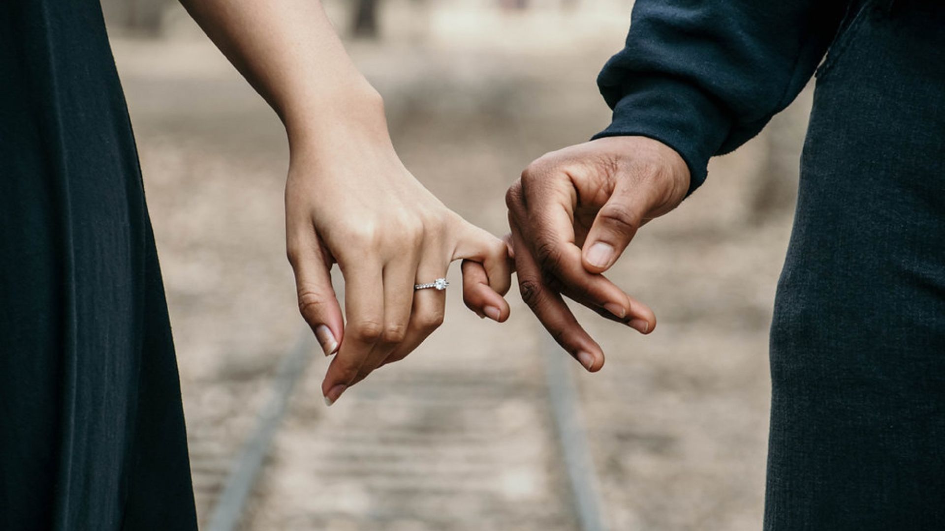 Imagem de um casal vestido com roupas pretas. Eles estão de mãos dadas, segurando os dedos.  Ela usa um lindo solitário.