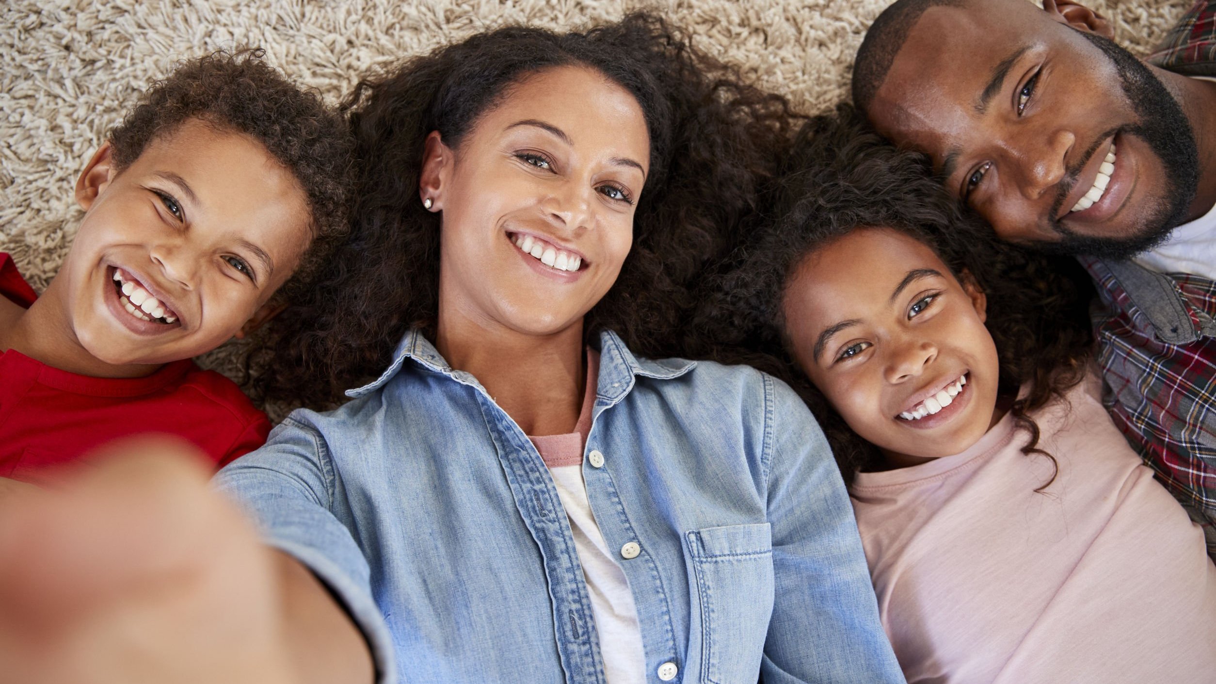 Família negra com mãe, pai, menino e menina sorrindo deitados num tapete bege.