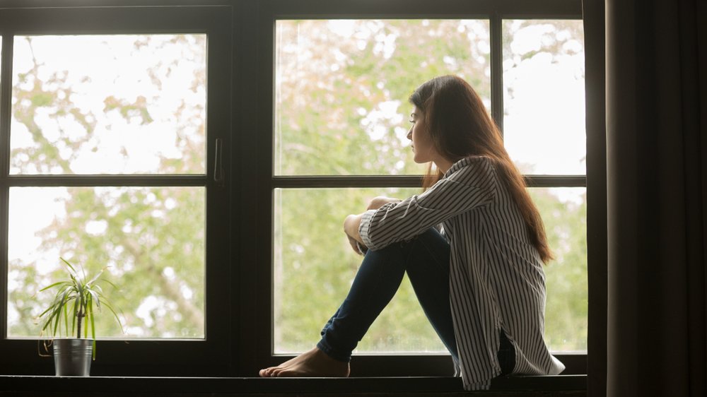 Jovem mulher sentada na janela, observando o lado de fora, pensativa.