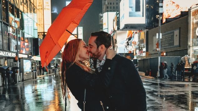 Casal de mulher e homem se beijando sob um guarda-chuva alaranjado. Eles estão em meio a uma rua de centro urbano.