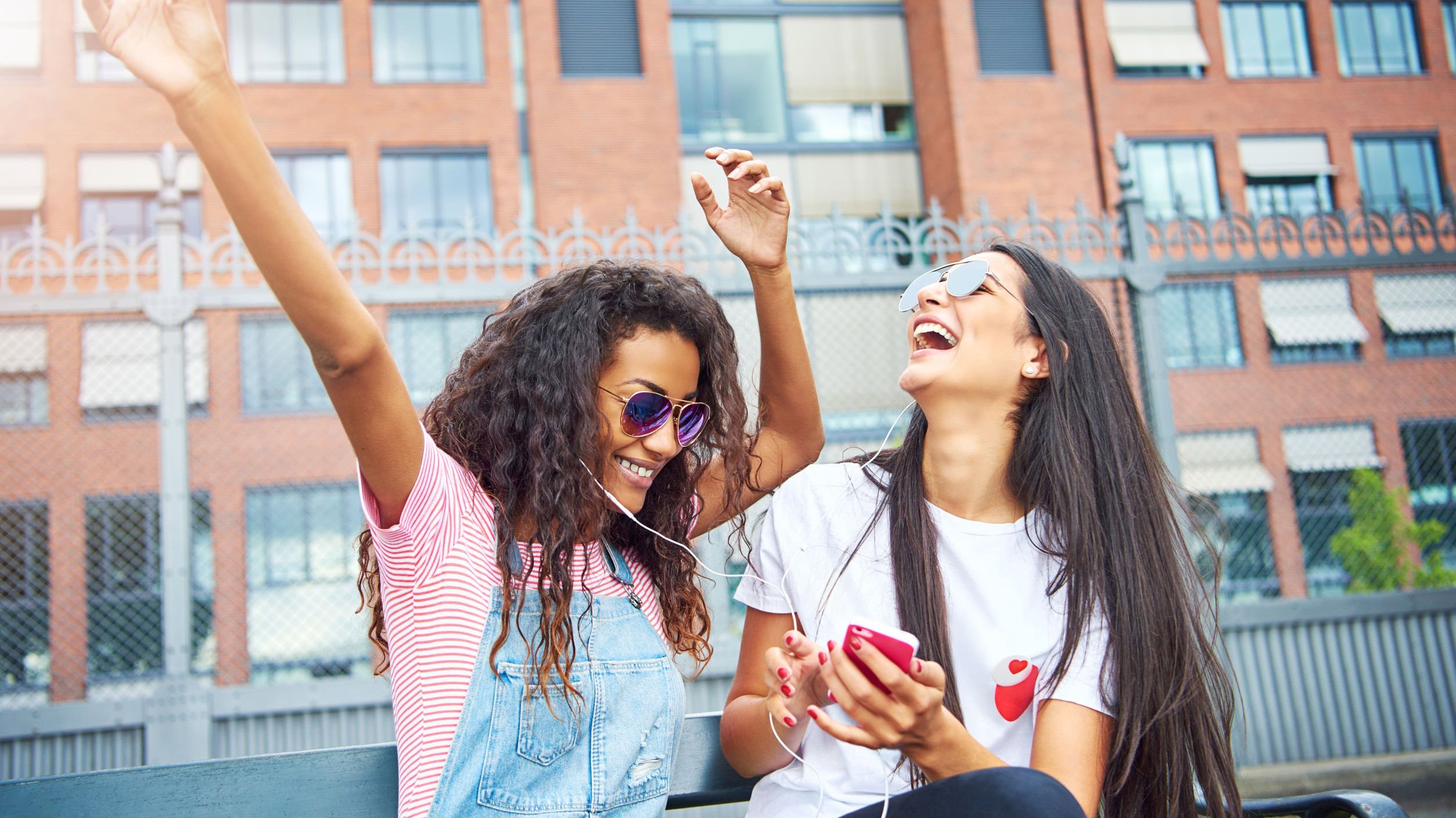 Duas jovens rindo, ouvindo música dividindo fone de ouvido. Uma segura o celular, e a outra tem os braços erguidos.