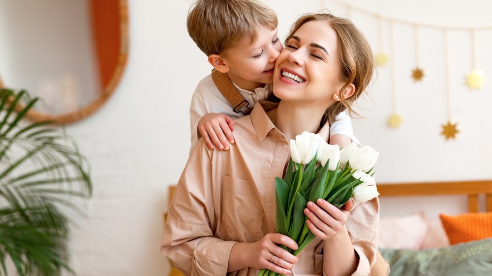 Menino beijando a mãe na bochecha enquanto ela segura um buquê de flores sorrindo