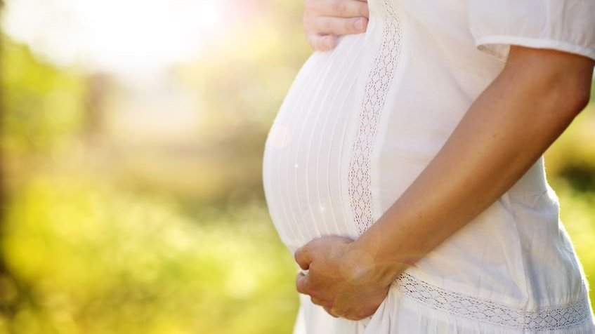 Mulher grávida usando vestido branco segura sua barriga com a mão esquerda embaixo e a mão direita em cima.