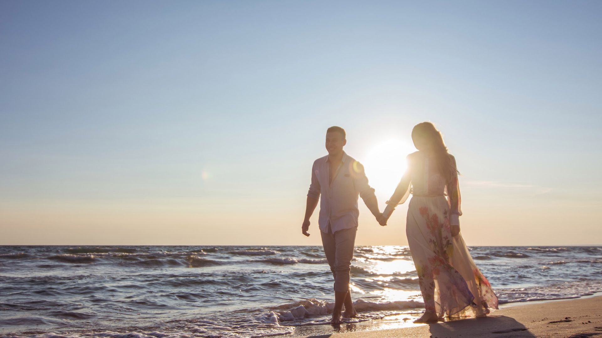 Imagem do mar com um casal de mãos dadas caminhando na areia.