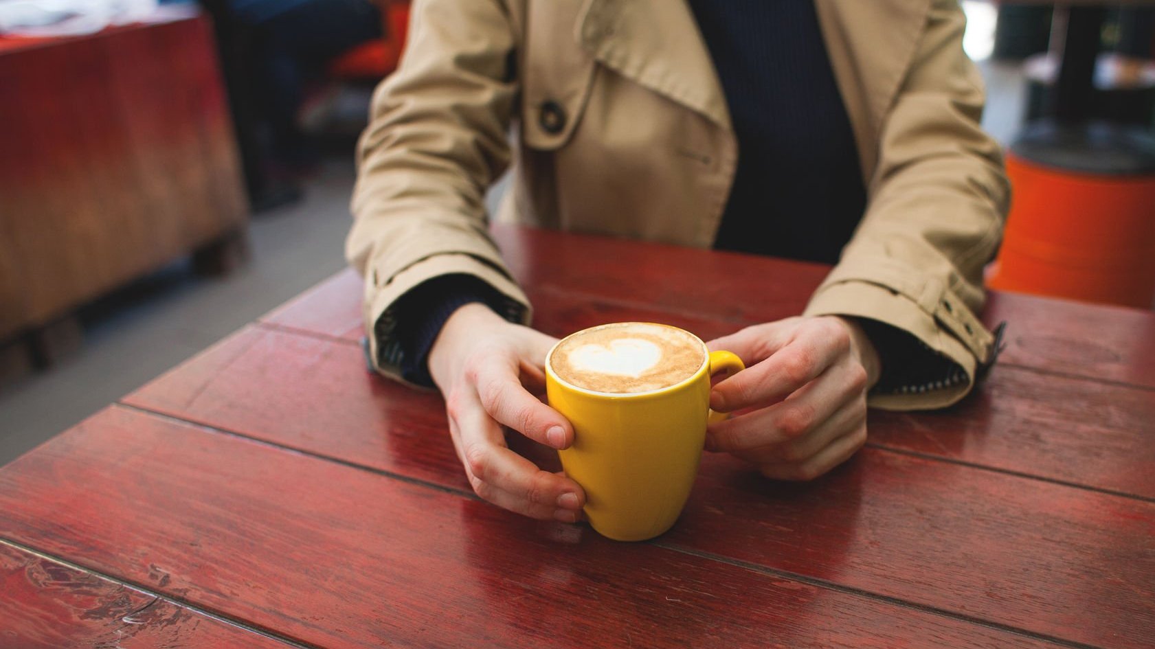 Pessoa vestindo casaco, sentada, apoiando os braços em uma mesa enquanto segura uma caneca com café com leite, e espuma em formato de coração.