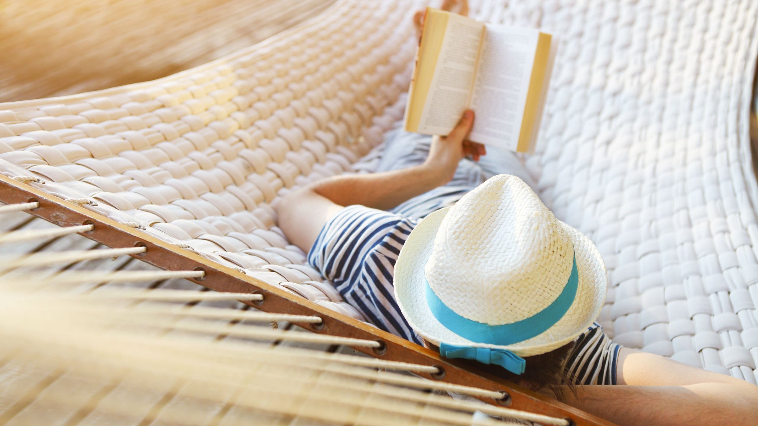 Homem branco deitado numa rede branca, usando chapéu e segurando um livro aberto.